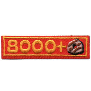 A rectangular red fabric patch with yellow embroidered text reading "7000+" and a small brown trefoil symbol on the right side.