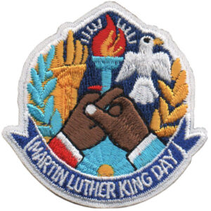 The MLK Patch (Copy) features a vibrant design of two hands—one light, one dark—joined in unity below a torch and a white dove, with "Martin Luther King Day" embroidered at the bottom.