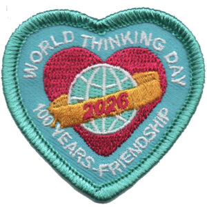 The 2025 World Thinking Day Patch (Copy) is a heart-shaped embroidered badge with a blue border, featuring a pink heart, globe, and yellow "2026" ribbon. The text reads "World Thinking Day, 100 Years Friendship.”.