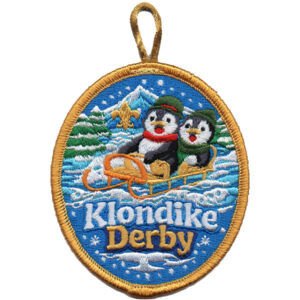 The Klondike Derby Penguins Patch (Copy) is an embroidered oval patch featuring two penguins on a sled, snowy trees and mountains in the background, “Klondike Derby” at the bottom, a gold border, and a hanging loop.