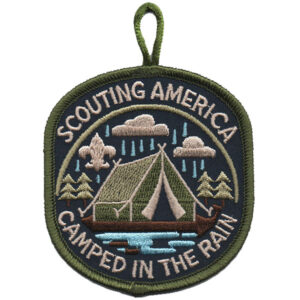 The Camped in the Rain Scouting America patch features a green tent, trees, rain, clouds, a lake, and a fleur-de-lis next to the tent. It has "Scouting America Camped in the Rain" text, a green border, and hanging loop.