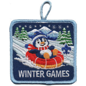 A square patch features a smiling penguin in a blue hat and scarf riding a red snow tube down a snowy hill with trees, mountains, and stars in the background. "CUB SCOUT GAMES" is written at the bottom.