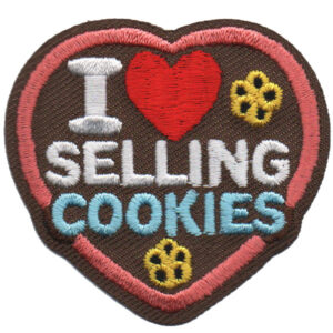 The I Love Selling Cookies Patch features a heart-shaped design with "I [red heart] SELLING COOKIES" embroidery, three yellow flower-shaped cookies, a brown background, and a pink border.